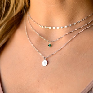 May include: Layered silver necklace with a small green gemstone pendant and a larger pendant with the letter "M" engraved on it.