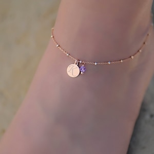 Puede incluir: Un delicado tobillo de oro rosa con un pequeño dije redondo grabado con la letra "K" y un acento de cristal morado.