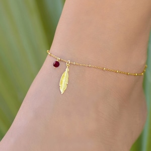 Peut inclure: Un bracelet de cheville en or avec une chaîne délicate et un petit charme en pierre rouge. Un charme en forme de plume dorée pend au charme en pierre.
