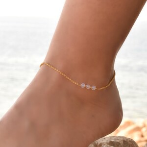 May include: A delicate gold anklet with three small white beads. The anklet is on a woman's ankle, with the background blurred.