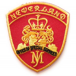 Peut inclure: Un écusson brodé rouge avec une bordure dorée et une couronne dorée. L'écusson porte l'inscription "Neverland" et "Office of Special Services" avec un "M" doré au centre.