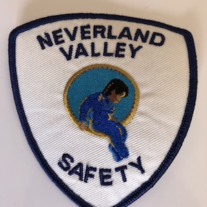Puede incluir: Parche blanco bordado con borde azul. El parche presenta el texto "Neverland Valley Safety" y un círculo azul y dorado con una figura de dibujos animados sentada dentro.