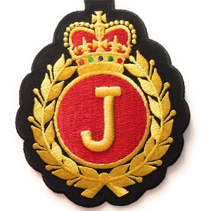 May include: A gold embroidered patch featuring a red circle with a gold letter J, encircled by a gold laurel wreath and topped with a gold crown.