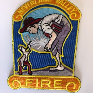 Peut inclure: Un écusson brodé avec une bordure jaune et les mots "Neverland Valley Fire" dans un design en forme de rouleau. L'écusson présente une image de dessin animé d'un homme avec un chapeau rouge et un petit chien.