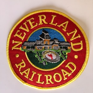 May include: Circular embroidered patch with a red border and the words "NEVERLAND RAILROAD" in yellow. The centre features a detailed illustration of a building and a circular emblem with a red "Y" on a green and blue background.