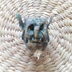 May include: A green and brown goblin face sculpture with a white beard made of straw. The sculpture is sitting on a woven straw mat.