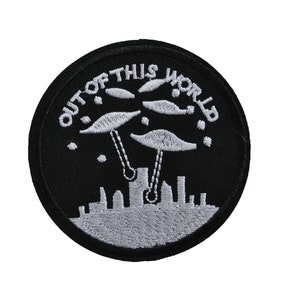 Peut inclure: Patch brodé noir et blanc avec une ville et trois soucoupes volantes. Le texte "OUT OF THIS WORLD" est écrit autour du bord du patch.