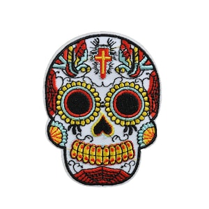 Peut inclure: Un crâne de sucre blanc avec des motifs floraux rouges, jaunes et bleus. Le crâne a une croix sur le front et un nez en forme de cœur.