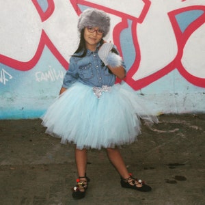 Pode incluir: Uma pessoa vestindo uma camisa jeans, uma saia de tule azul claro e sapatos pretos com detalhes de pompom. Eles também estão usando um chapéu de pele cinza, óculos e uma luva de pele combinando. O fundo apresenta arte de grafite.