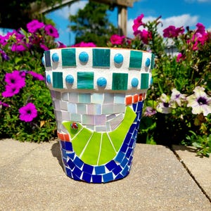 May include: A mosaic-tiled flower pot with a bird design. The pot features a white rim with turquoise and teal accents. The bird is green with a red eye, set against a blue and white background. The pot is outdoors, surrounded by flowers.