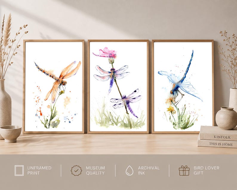 May include: A set of three vertical wall art prints featuring watercolour illustrations of dragonflies. The left print shows an orange dragonfly, the centre print displays two purple dragonflies on a stem, and the right print depicts a blue dragonfly.