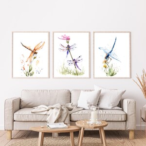 May include: A set of three framed wall art prints featuring watercolour illustrations of dragonflies. The left print shows an orange dragonfly, the centre print displays two purple dragonflies on a plant, and the right print depicts a blue dragonfly.