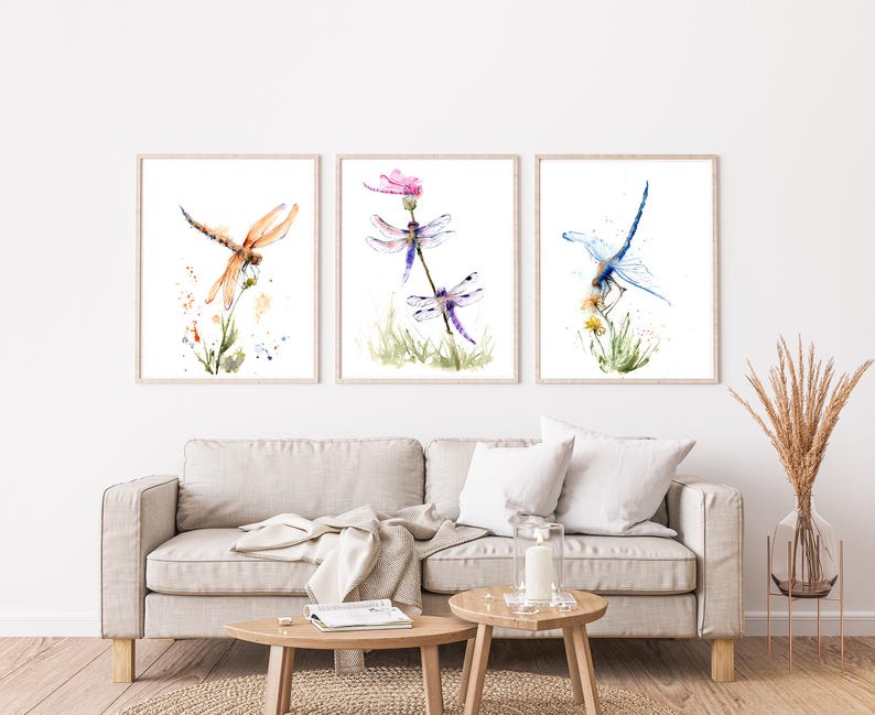 May include: A set of three framed wall art prints featuring watercolour illustrations of dragonflies. The left print shows an orange dragonfly, the centre print displays two purple dragonflies on a plant, and the right print depicts a blue dragonfly.