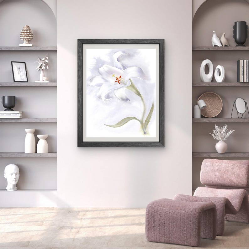 May include: A framed watercolor painting of a white lily with a green stem and leaves. The artwork is set against a soft, light background and framed in a dark gray wooden frame. The painting is displayed on a white wall.