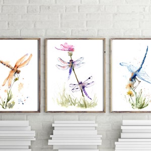 May include: A set of three framed watercolour prints featuring dragonflies. The left print shows an orange dragonfly on a plant. The centre print displays two purple dragonflies and a pink flower. The right print depicts a blue dragonfly on yellow flowers.