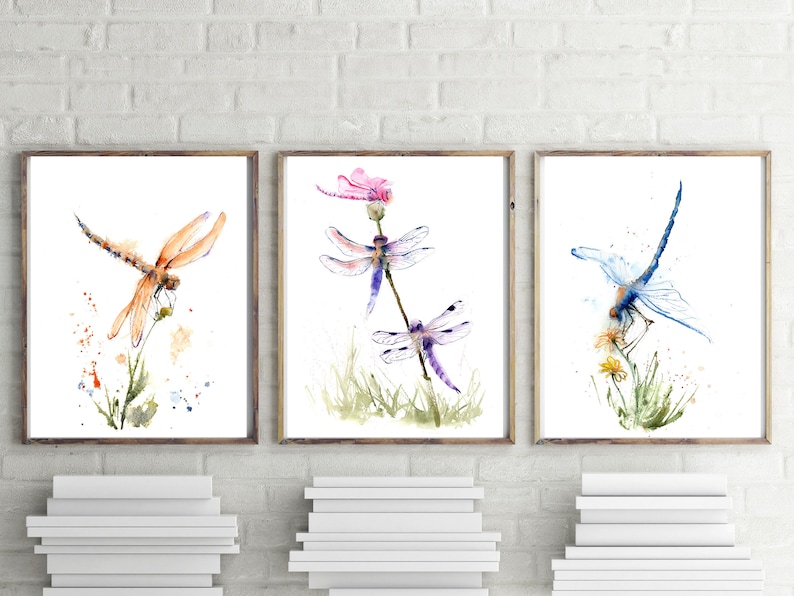May include: A set of three framed watercolour prints featuring dragonflies. The left print shows an orange dragonfly on a plant. The centre print displays two purple dragonflies and a pink flower. The right print depicts a blue dragonfly on yellow flowers.