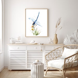 May include: A framed watercolour print featuring a blue dragonfly perched on yellow flowers and green grass. The artwork is displayed in a light wood frame, showcasing detailed brushstrokes and vibrant blue, yellow, and green paint splatters.