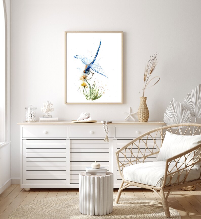 May include: A framed watercolour print featuring a blue dragonfly perched on yellow flowers and green grass. The artwork is displayed in a light wood frame, showcasing detailed brushstrokes and vibrant blue, yellow, and green paint splatters.