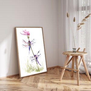 May include: A framed watercolour print featuring two dragonflies with purple and blue wings resting on a thin stem, with a pink flower at the top and green grass at the base, all set against a clean white background within a light wood frame.