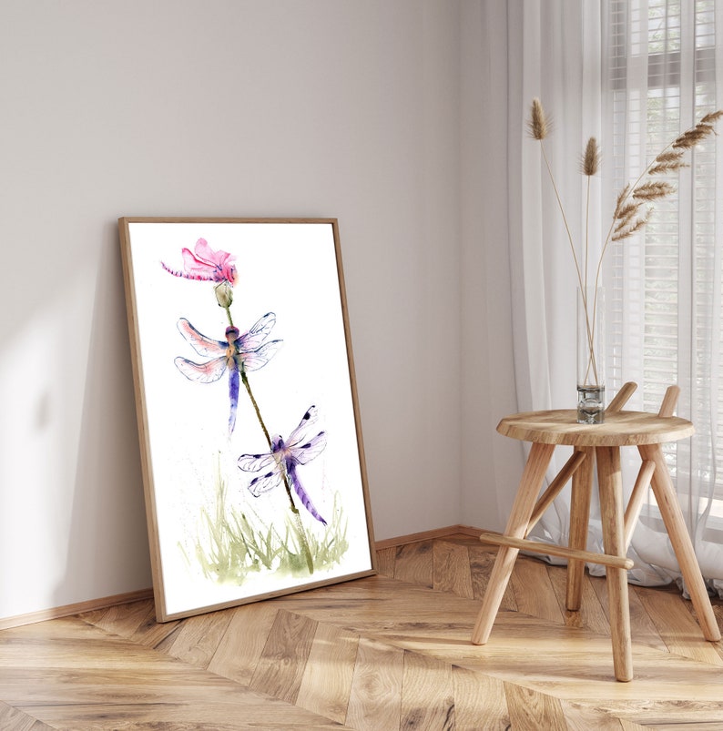 May include: A framed watercolour print featuring two dragonflies with purple and blue wings resting on a thin stem, with a pink flower at the top and green grass at the base, all set against a clean white background within a light wood frame.