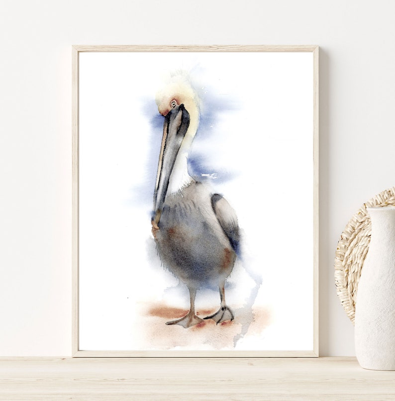May include: Watercolor painting of a brown pelican standing on a sandy beach with a blue and white watercolor background.