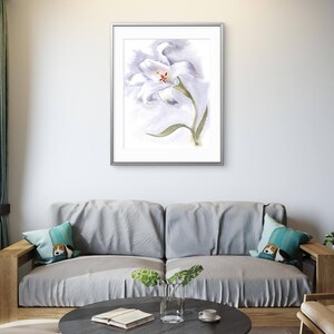 May include: A framed watercolor painting of a white lily with a yellow center and green stem. The artwork is displayed above a gray sofa with decorative pillows. A round, dark gray coffee table with a book, vase, and cup is in front of the sofa.