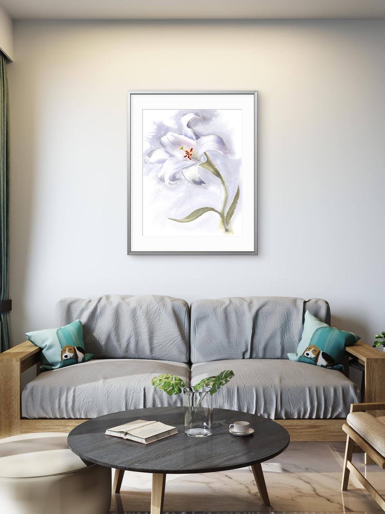 May include: A framed watercolor painting of a white lily with a yellow center and green stem. The artwork is displayed above a gray sofa with decorative pillows. A round, dark gray coffee table with a book, vase, and cup is in front of the sofa.