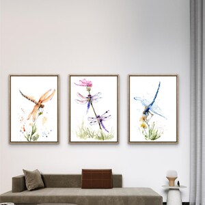 May include: A set of three framed wall art prints featuring watercolour illustrations of dragonflies. The left print shows an orange dragonfly, the centre print displays two purple dragonflies on a flower, and the right print depicts a blue dragonfly.