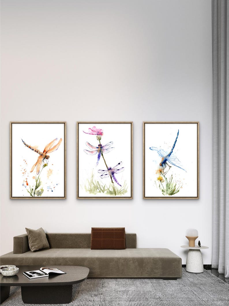 May include: A set of three framed wall art prints featuring watercolour illustrations of dragonflies. The left print shows an orange dragonfly, the centre print displays two purple dragonflies on a flower, and the right print depicts a blue dragonfly.