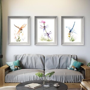 May include: Three framed watercolour prints displayed in a row, each featuring a dragonfly perched on a plant. The left print shows an orange dragonfly, the centre print depicts two purple dragonflies, and the right print features a blue dragonfly.
