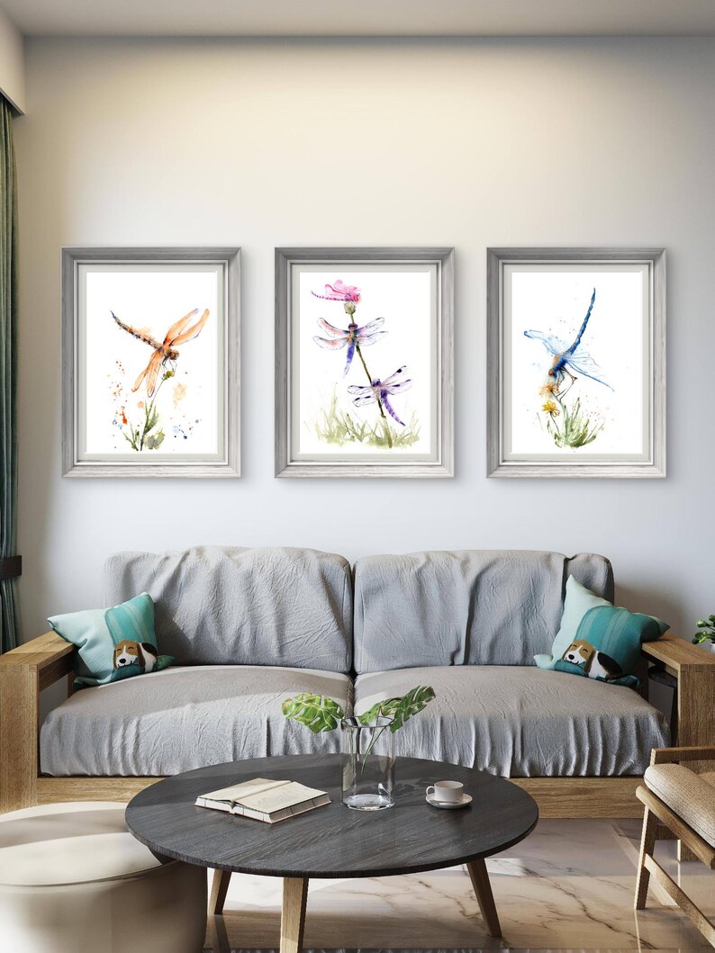 May include: Three framed watercolour prints displayed in a row, each featuring a dragonfly perched on a plant. The left print shows an orange dragonfly, the centre print depicts two purple dragonflies, and the right print features a blue dragonfly.