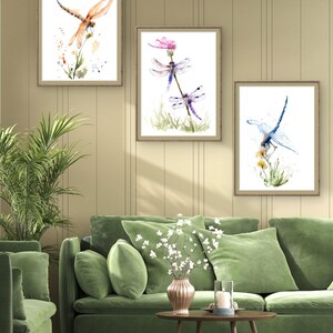 May include: Three framed wall art prints featuring watercolour illustrations of dragonflies. The left print shows an orange dragonfly, the centre print displays two purple dragonflies, and the right print depicts a blue dragonfly, all perched on stems.