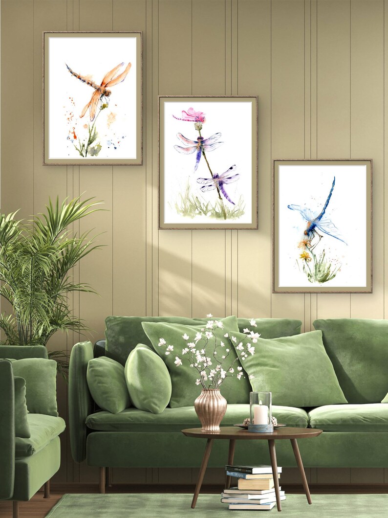 May include: Three framed wall art prints featuring watercolour illustrations of dragonflies. The left print shows an orange dragonfly, the centre print displays two purple dragonflies, and the right print depicts a blue dragonfly, all perched on stems.