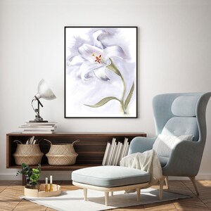 May include: A framed watercolor painting of a white lily with a green stem and leaf. The artwork is displayed in a living room setting, with a light blue armchair, ottoman, and wooden cabinet.
