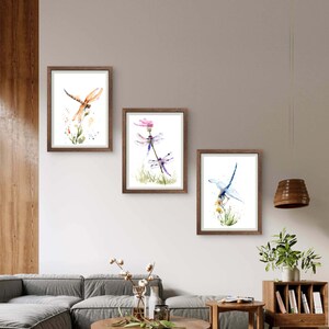 May include: Three framed watercolour prints featuring dragonfly illustrations. The left print shows an orange dragonfly, the centre print displays two purple dragonflies on a flower, and the right print depicts a blue dragonfly perched on yellow flowers.