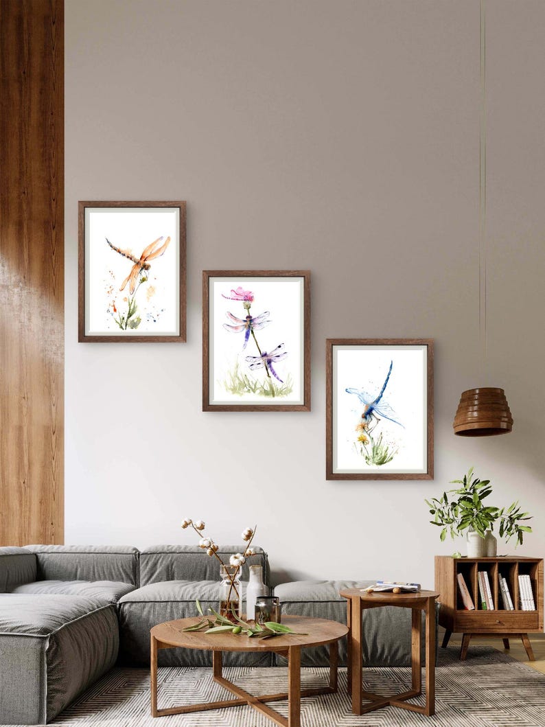 May include: Three framed watercolour prints featuring dragonfly illustrations. The left print shows an orange dragonfly, the centre print displays two purple dragonflies on a flower, and the right print depicts a blue dragonfly perched on yellow flowers.
