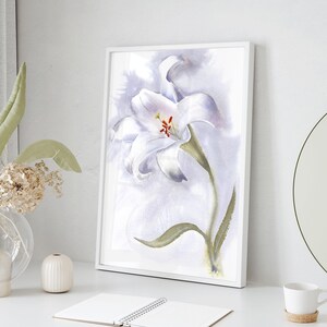 May include: A framed watercolor painting of a white lily with green leaves and a red center. The artwork is displayed on a white surface, next to a vase with dried flowers and a small white ceramic vase.