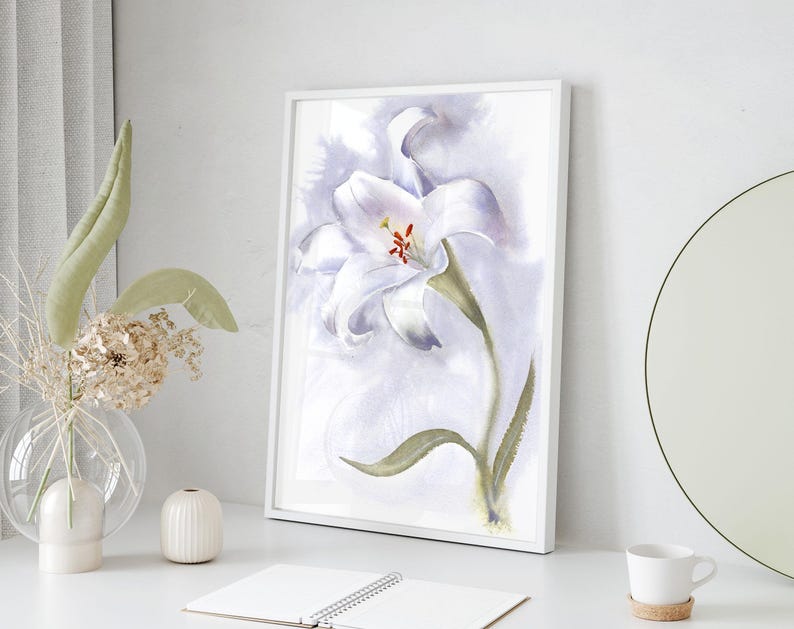 May include: A framed watercolor painting of a white lily with green leaves and a red center. The artwork is displayed on a white surface, next to a vase with dried flowers and a small white ceramic vase.