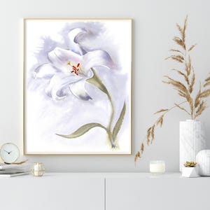 May include: Watercolor painting of a white lily with a green stem and leaves, set against a soft, lavender-toned background. The artwork is framed in a gold-colored frame. The painting is displayed on a white cabinet, with a clock, candle, and vase.