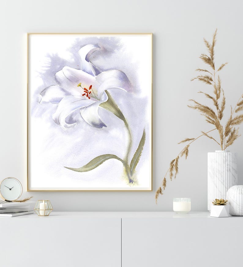 May include: Watercolor painting of a white lily with a green stem and leaves, set against a soft, lavender-toned background. The artwork is framed in a gold-colored frame. The painting is displayed on a white cabinet, with a clock, candle, and vase.