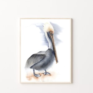 May include: Watercolor painting of a brown pelican standing on a sandy beach. The pelican has a white head and a long, orange beak. The background is a soft blue watercolor wash.