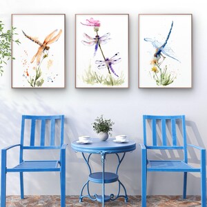 May include: Three framed watercolour prints featuring dragonflies in orange, purple, and blue hues, displayed above two blue metal chairs and a matching round bistro table with two white cups and a small potted plant on a stone patio.