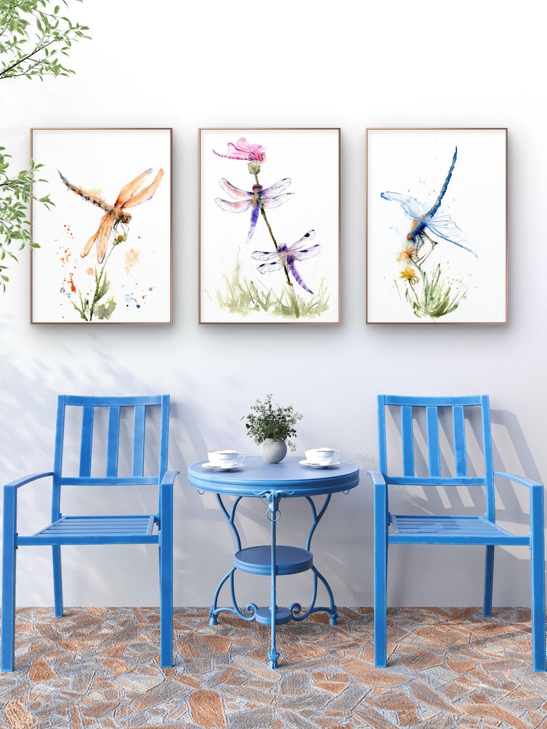 May include: Three framed watercolour prints featuring dragonflies in orange, purple, and blue hues, displayed above two blue metal chairs and a matching round bistro table with two white cups and a small potted plant on a stone patio.