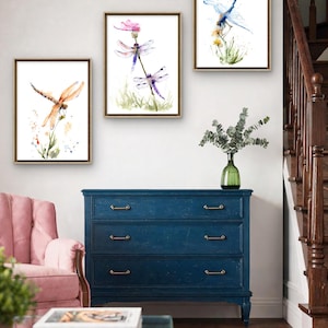 May include: Three framed watercolour prints featuring dragonflies in flight and perched on plants. The left print shows an orange dragonfly, the centre print displays two purple dragonflies on a flower, and the right print depicts a blue dragonfly.