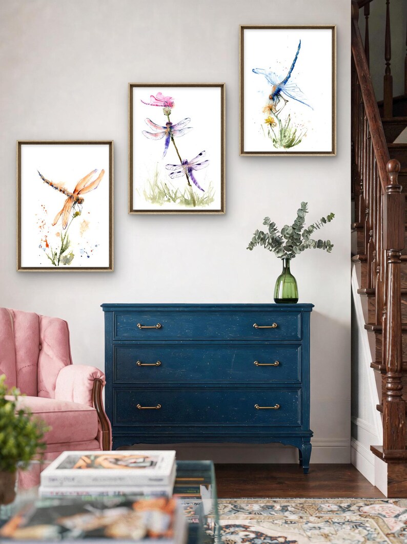 May include: Three framed watercolour prints featuring dragonflies in flight and perched on plants. The left print shows an orange dragonfly, the centre print displays two purple dragonflies on a flower, and the right print depicts a blue dragonfly.