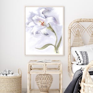 May include: Watercolor painting of a white lily flower with green stem and leaves, set against a soft, light purple background. The artwork is framed in a light wood frame and hangs above a wicker nightstand and basket.