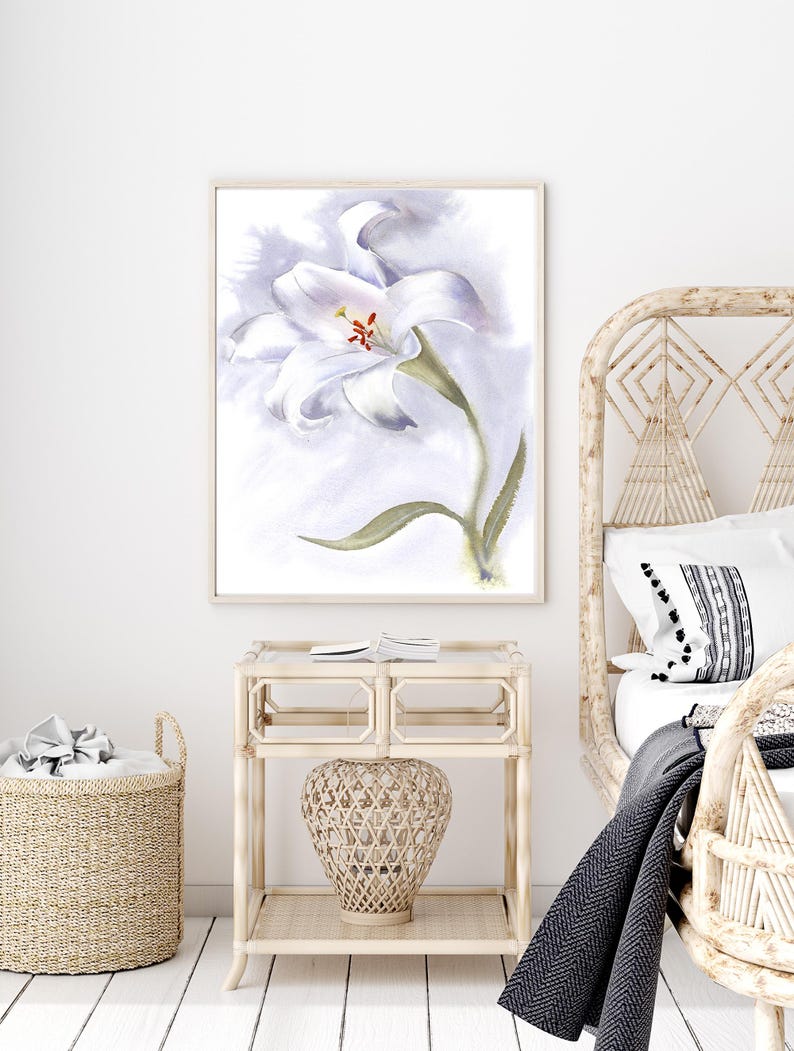 May include: Watercolor painting of a white lily flower with green stem and leaves, set against a soft, light purple background. The artwork is framed in a light wood frame and hangs above a wicker nightstand and basket.