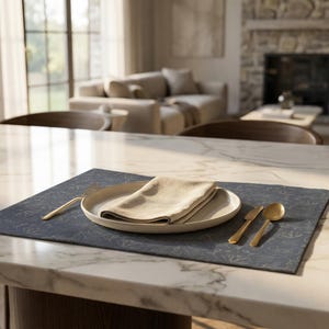 Può includere: Un'apparecchiatura da tavola con piatto color crema, tovagliolo beige piegato e posate dorate. L'apparecchiatura poggia su un sottopiatto blu scuro con un sottile motivo floreale. La tavola è in una stanza ben illuminata.