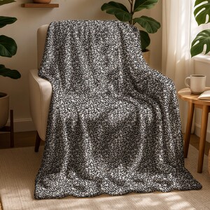 May include: A soft, gray and white throw blanket with a leopard print pattern is draped over a beige armchair. The blanket's design features small, irregular spots, creating a textured appearance. The blanket is made of a soft material.