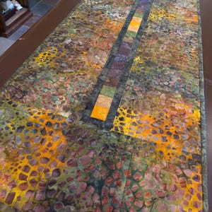 May include: A rectangular table runner with a vibrant, abstract design. The runner features a patchwork of colors including yellow, green, brown, and purple. A central stripe of alternating colored squares runs down the center. The runner is likely made of fabric.